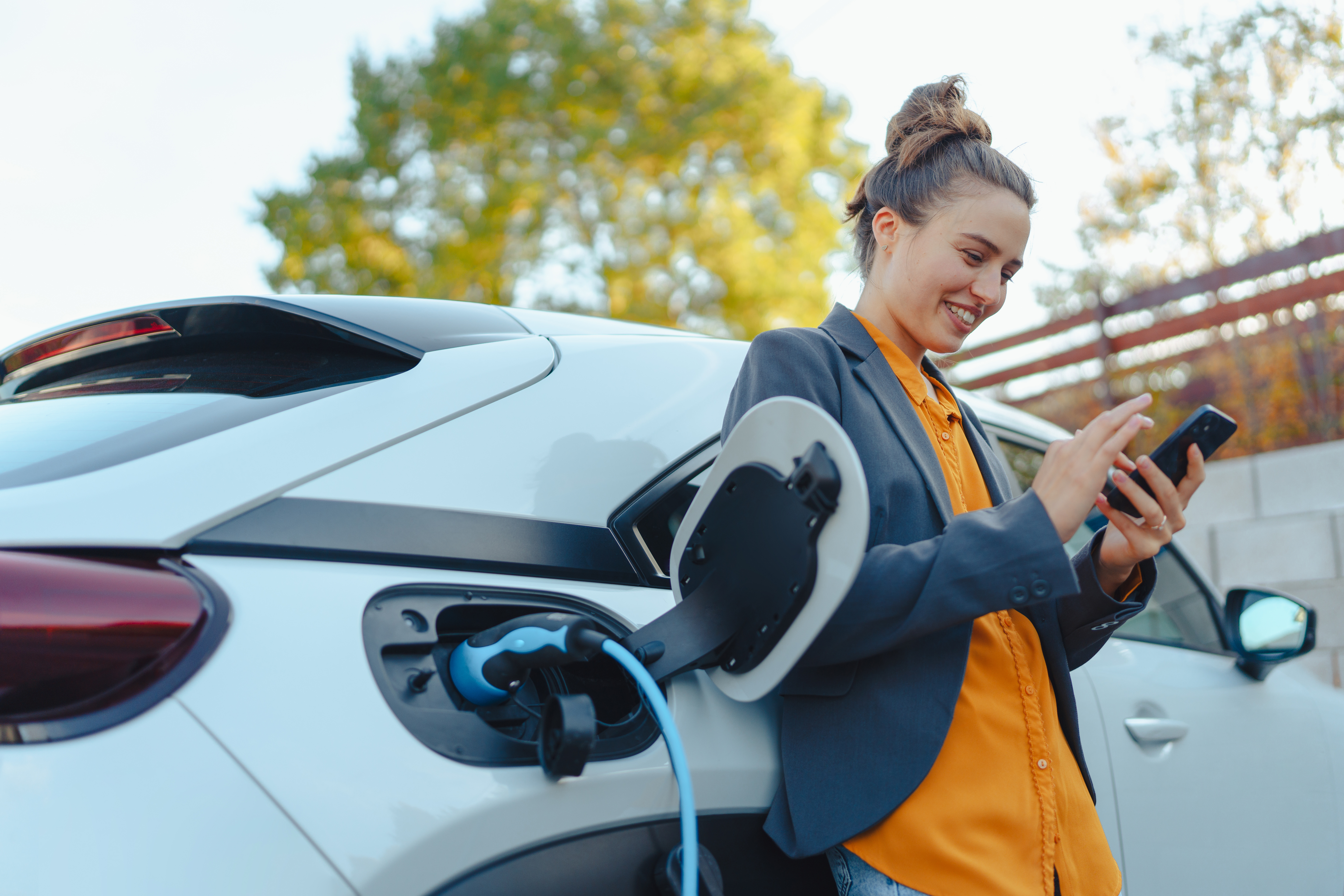 Young woman with smartphone waiting while electric car charging in home charging station, sustainable and economic transportation concept.